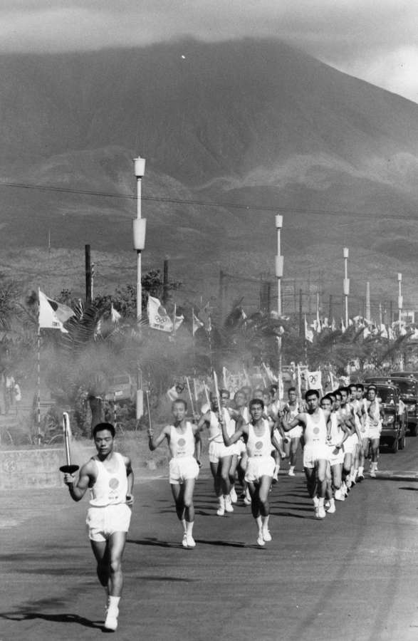 桜島をバックにトーチを掲げ、力強く駆ける東京五輪の聖火リレー。走者は平川拓也さん＝1964年9月9日、鹿児島市上之園町のナポリ通り