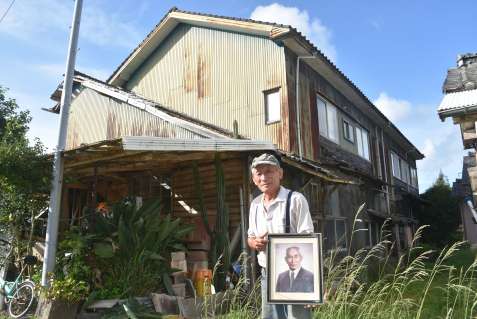 旧阿久根小校舎の前で、父横峯勉さんの遺影を持つ均さん＝出水市高尾野