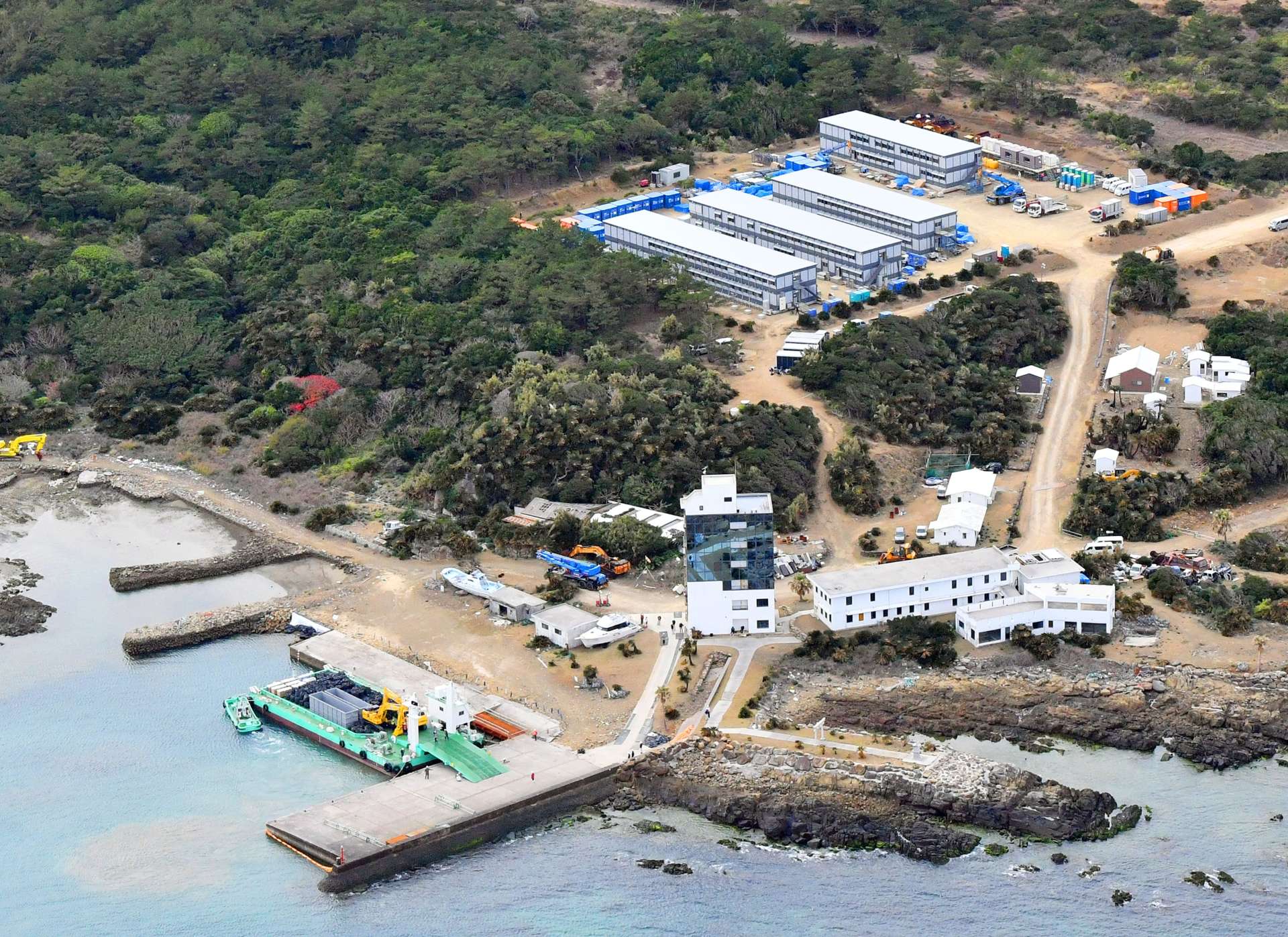 【別カット】重機や資材を積んだ台船が接岸し、工事の準備が進む葉山港周辺。上は新設された作業員用宿舎＝１２日午後２時半、西之表市の馬毛島（本社チャーター機から撮影）