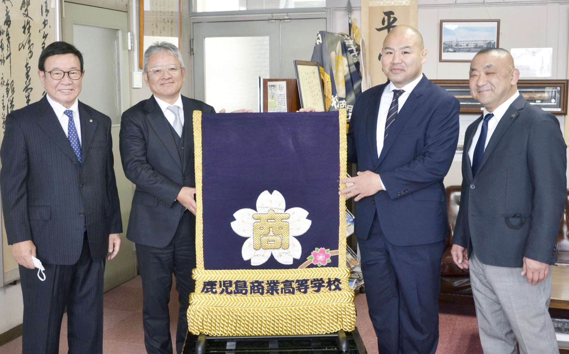 化粧まわしを返還した里山浩作さん（右から２人目）と受け取った鹿児島商業高校の堀之内尚郎校長（同３人目）＝２４日、鹿児島市西坂元町の同校