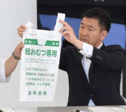 志布志市の紙おむつ専用指定ごみ袋を持つ柳本顕環境政務官（中央）＝１０日、大崎町菱田のそおリサイクルセンター