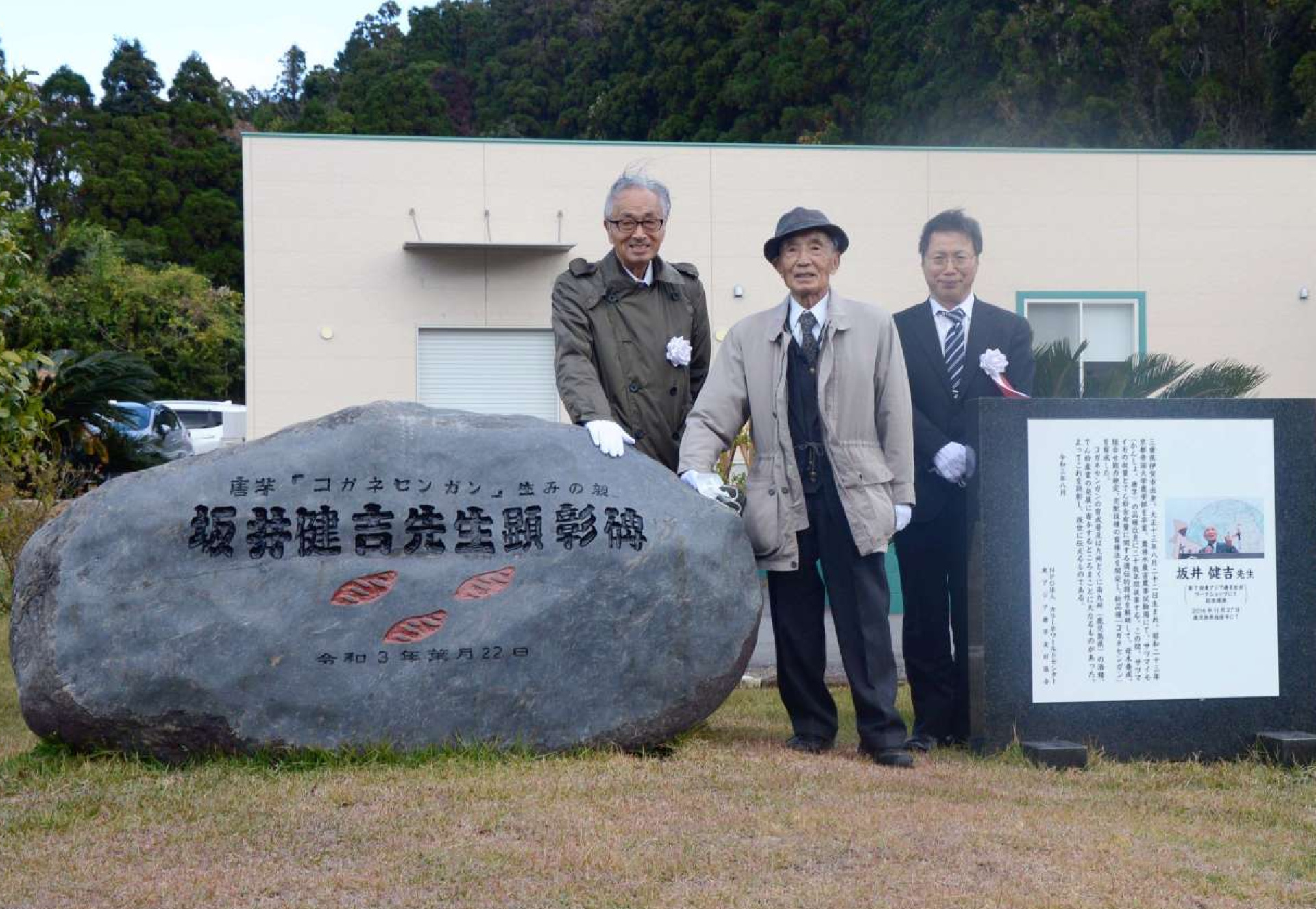 ２０２１年１２月、コガネセンガンの生みの親をたたえる顕彰碑の横に立つ坂井健吉さん（中央）＝鹿屋市