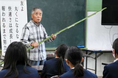 手作りの竹やりを手に戦時中の暮らしを紹介する政田一雄さん＝中種子町の増田小学校