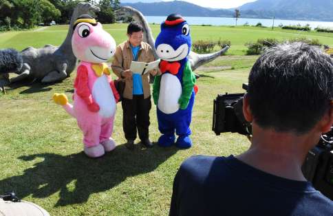 池田湖や開聞岳をバックに、地元のマスコットキャラクターと撮影に臨む主演の内藤剛志さん＝９月、指宿市のえぷろんはうす池田