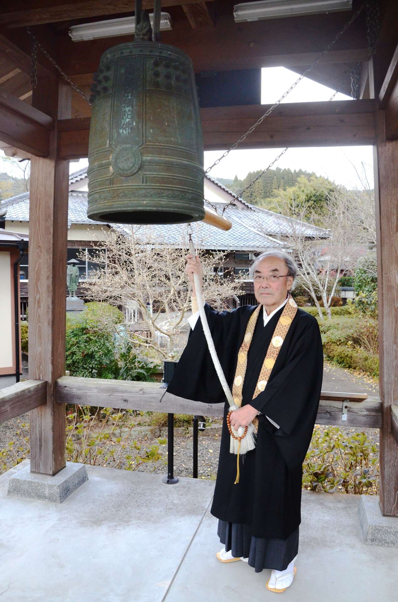 泉福寺の鐘楼につるされた小型の半鐘を、「恒久平和を願ってつく」と語る横井晃仁住職＝さつま町永野