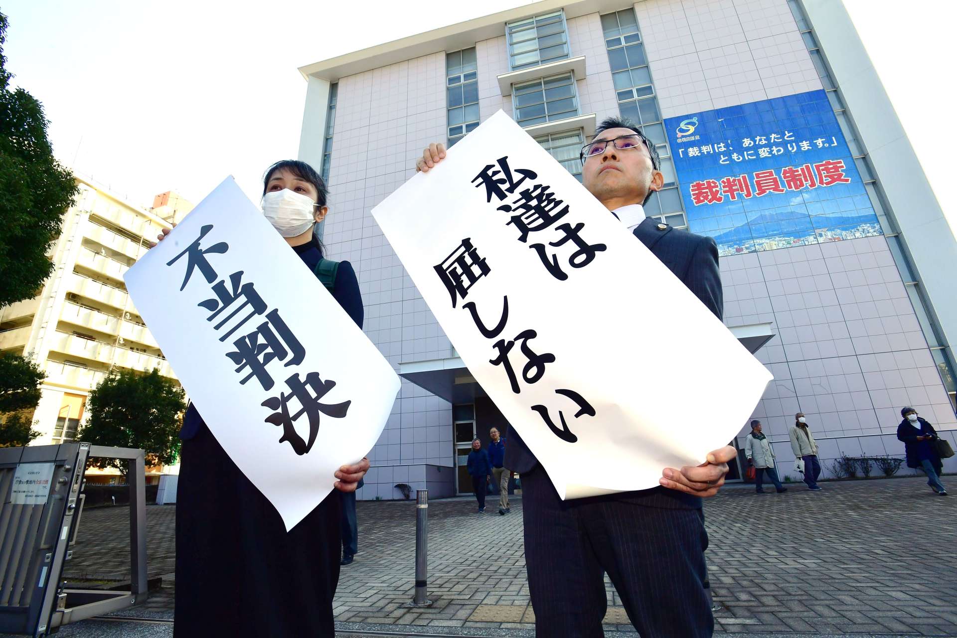「不当判決」の紙を掲げる森永明子原告団長（左）ら＝２１日午後３時９分、鹿児島市山下町