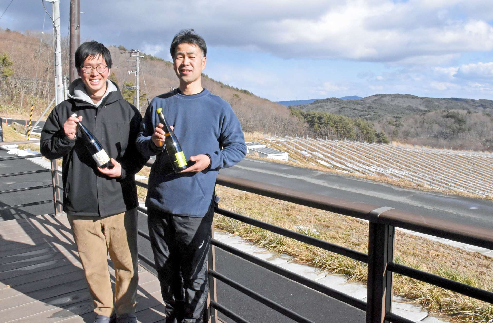 ブドウ畑の前でかわうちワインを手にする安達貴さん（左）と遠藤一美さん＝２月、福島県川内村のかわうちワイナリー