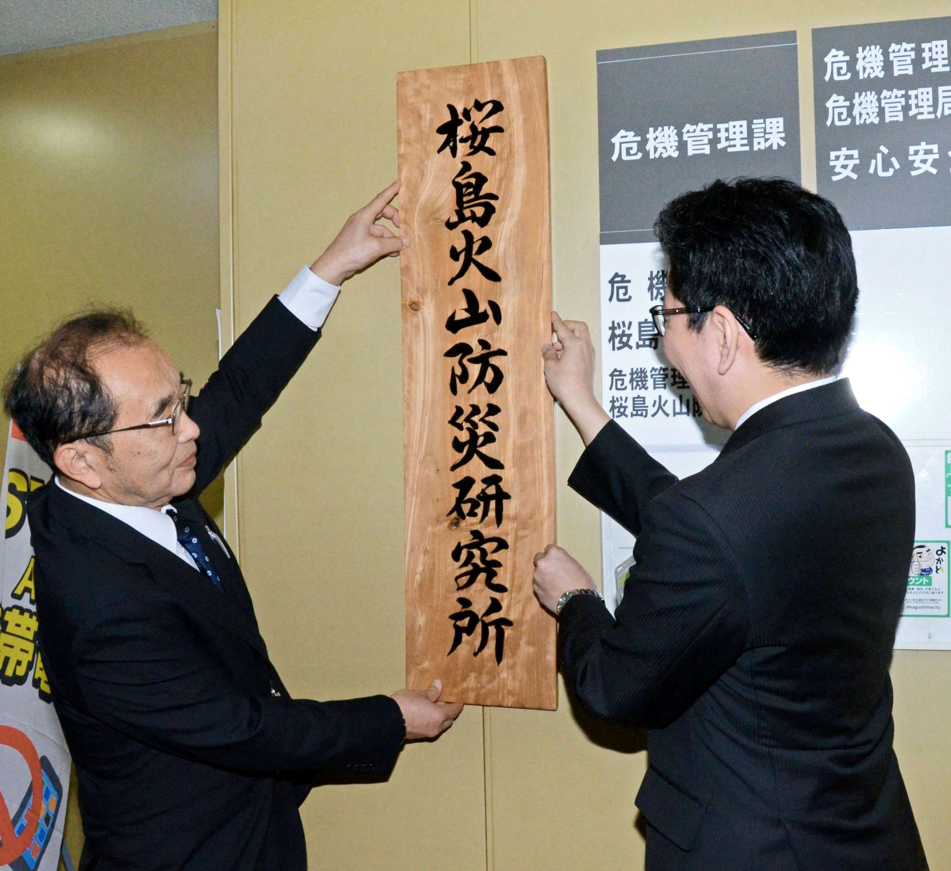 １日発足した「桜島火山防災研究所」の看板を掛ける井口正人所長（左）と下鶴隆央市長＝１日、鹿児島市役所