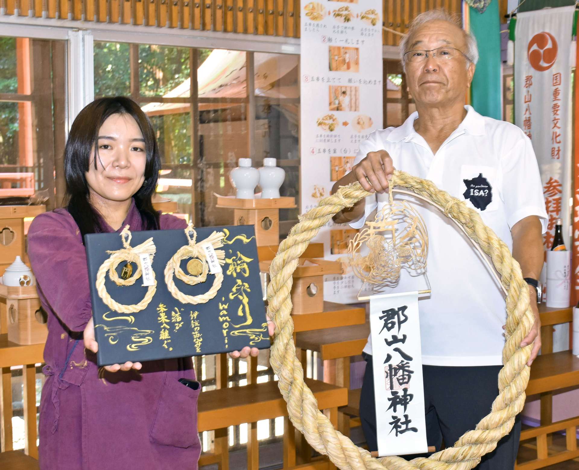 大阪・関西万博に展示し、郡山八幡神社に奉納された「輪守り」を持つ山下和弘氏子総代会長（右）と販売用を持つ和繼の井料明歩代表（左）＝伊佐市大口大田