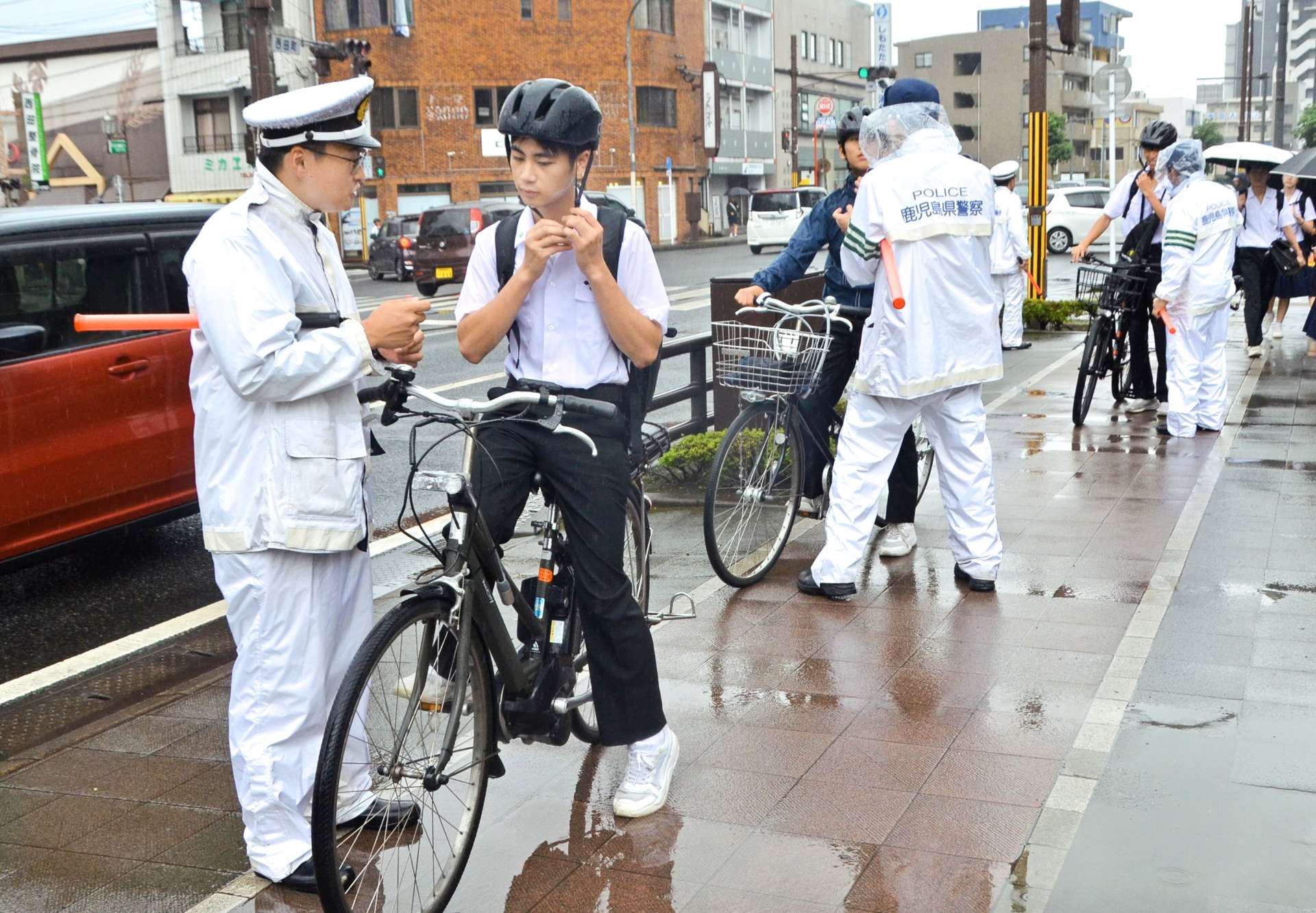 ヘルメットのあごひもを締めるよう生徒に指導する警察官＝１４日午前８時前、鹿児島市西田３丁目