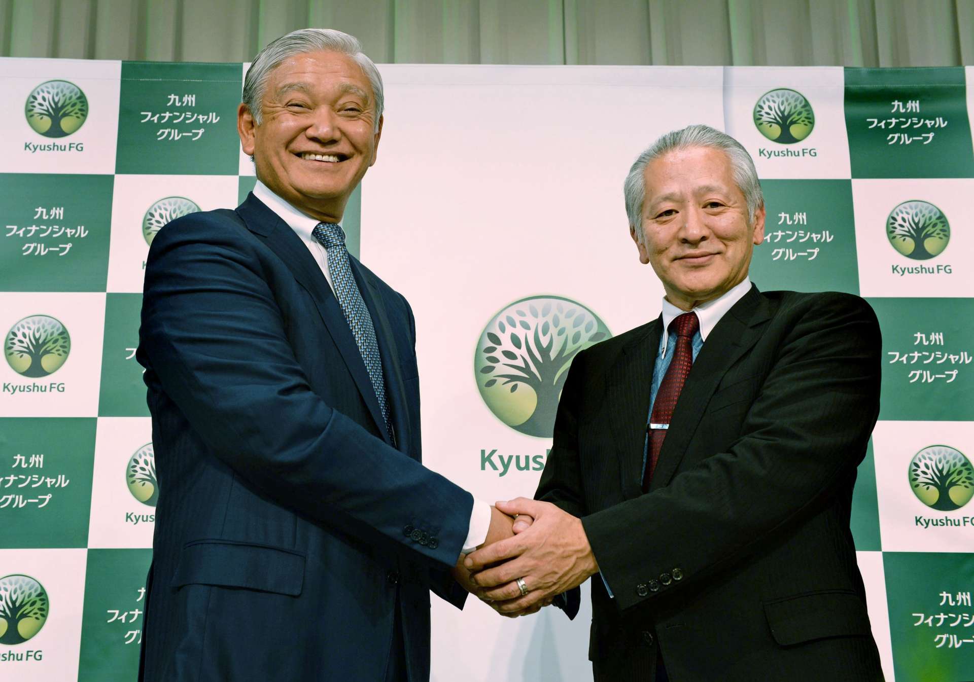 九州フィナンシャルグループ設立で握手を交わす肥後銀行の甲斐隆博頭取（左）と鹿児島銀行の上村基宏頭取（肩書きはいずれも当時）＝２０１５年１０月１日、熊本市中央区