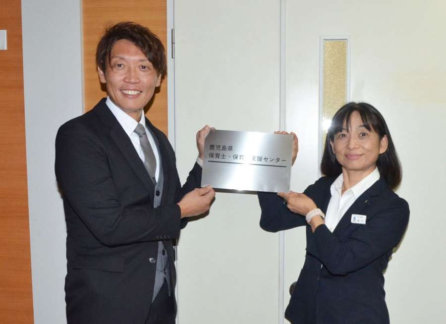 センター名が入ったプレートを持つ県子ども政策局の新川康枝局長（右）と県保育連合会の帯田英児会長＝１日、鹿児島市の県老人福祉会館