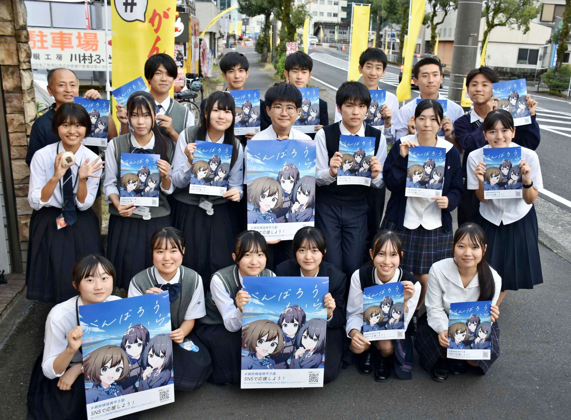 黄色い旗が並ぶ通りで、「がんばろうあいら」のポスターを持つ高校生ら＝姶良市加治木