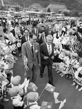 昭和５９年１０月２５日、園児らのふる旗に出迎えられるクイーンズランド州の首相一行。夕刊の紙面では「鹿児島に到着した２匹のコアラはＶＩＰ並みの待遇。ものものしい輸送作戦で空港から平川動物園まで運ばれた。特別仕様の専用車は、パンダ輸送にも使われたもので、パトカー先導も天皇陛下の来鹿の際と同じ体制だったという。平川動物公園では保育園児らが熱烈な歓迎。それにこたえて２匹が公開されると歓声があがった」と報じている。