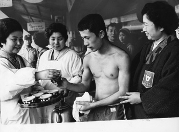 昭和４４年２月１８日、宮之城中継点で健闘した大島チームの選手をいたわる地元婦人会の方々。紙面では「県下一周駅伝第３日のフィナーレ。遠来の大島チームに寄せる温かい声援はこの日だけでなく、コースのあちこちで選手たちを励まし続けている。（中略）宮之城中継点では走り込んできた初出場の屋信考選手を同町の婦人会員が、わが子をだきかかえるようにして力走をたたえ、暖かいお茶で元気づけていた」と伝えている。