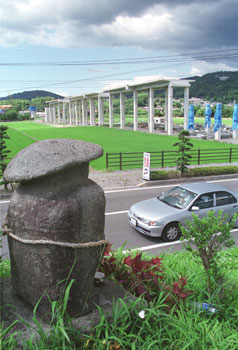 平成１２年８月、出水市街地で進む九州新幹線鹿児島ルートの工事を見守る田の神様。「５日にあった出水駅高架橋の安全祈願祭で、同市を通る全区間が着工。明治９年から出水平野を見守り続けた田の神様には、高架橋を疾走する新幹線はどう映る？」と紙面。