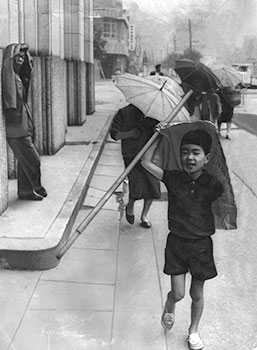昭和４３年５月２５日、鹿児島市役所前、町並みがかすむほどの灰をかさや紙切れでよける人たち。紙面では「まるで砂ばくのあらしのよう、先が見えないほど。婦人たちは日がさをさして灰を防ごうとしたが、風にあおられて舞い上がるので効果なくすっかり灰まみれ。子供たちは悲鳴をあげて近くの建て物に避難、単車乗りは目があけられず運転ストップ、四輪車もノロノロ運転と、大きな火ばちをさかさにしたような騒ぎだった」と報じている。